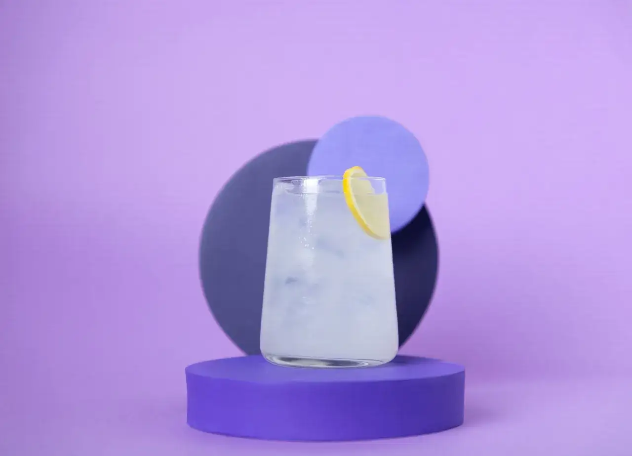 Erfrischendes bitter lemon tonic water mit Eis und Zitronenscheibe auf lila Podest vor abstrakten Kreisen.
