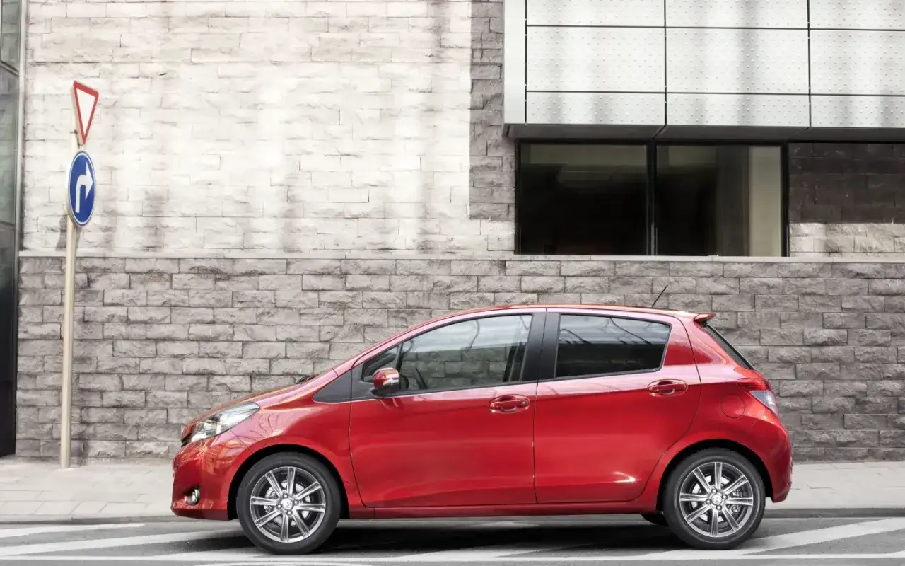 Czerwony Toyota Yaris Sedan zaparkowany przy nowoczesnym budynku z kamienną fasadą.