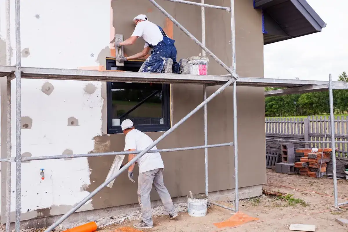 Zwei Handwerker verputzen eine Hausfassade. Die Ger&uuml;ste und Materialien deuten auf die bevorstehenden Fassade verputzen Kosten hin.