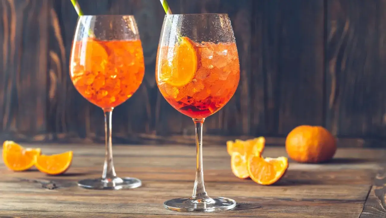 Tv&aring; glas med Aperol Spritz, is och apelsinskivor, p&aring; ett tr&auml;bord med apelsinklyftor och en hel apelsin.
