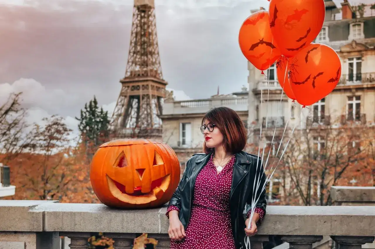 Une femme avec des ballons orange et une citrouille sculptée, devant la Tour Eiffel. Idée de quoi faire pour Halloween à Paris.