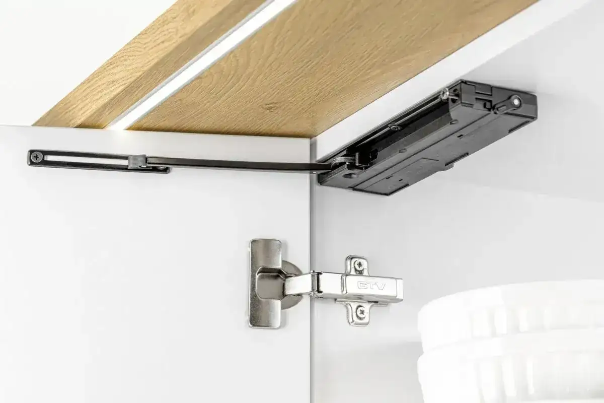 Mechanizm do szafek kuchennych, pokazujący jak zamontować uchwyty do szafek IKEA. Widoczny zawias i siłownik gazowy.