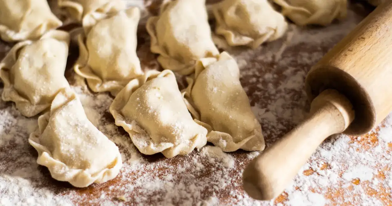 Surowe pierogi ruskie leżą na drewnianej desce, obok wałek do ciasta.