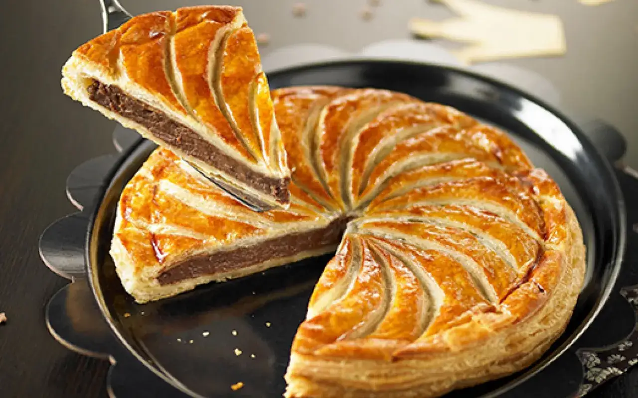Une part de galette des rois pépite de chocolat est soulevée, révélant sa garniture gourmande. La pâte feuilletée dorée est joliment décorée.