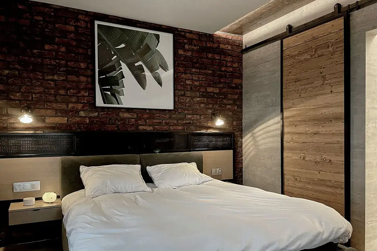 Sypialnia w stylu loft z ceglaną ścianą, dużym ł&oacute;żkiem i drewnianymi drzwiami przesuwnymi.