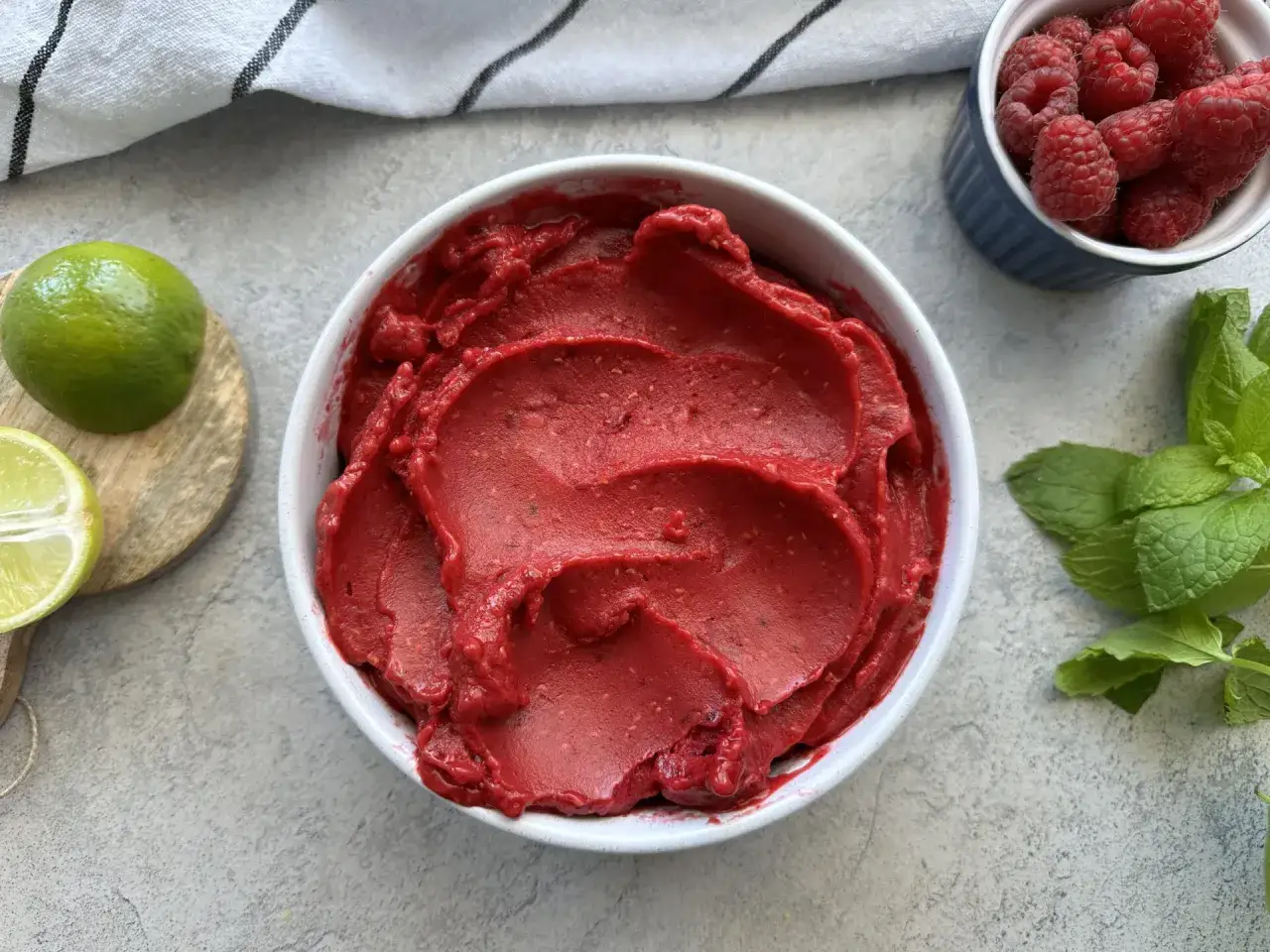 Jak zrobić sorbet malinowy? W misce pyszny, czerwony sorbet, obok maliny, limonka i mięta.
