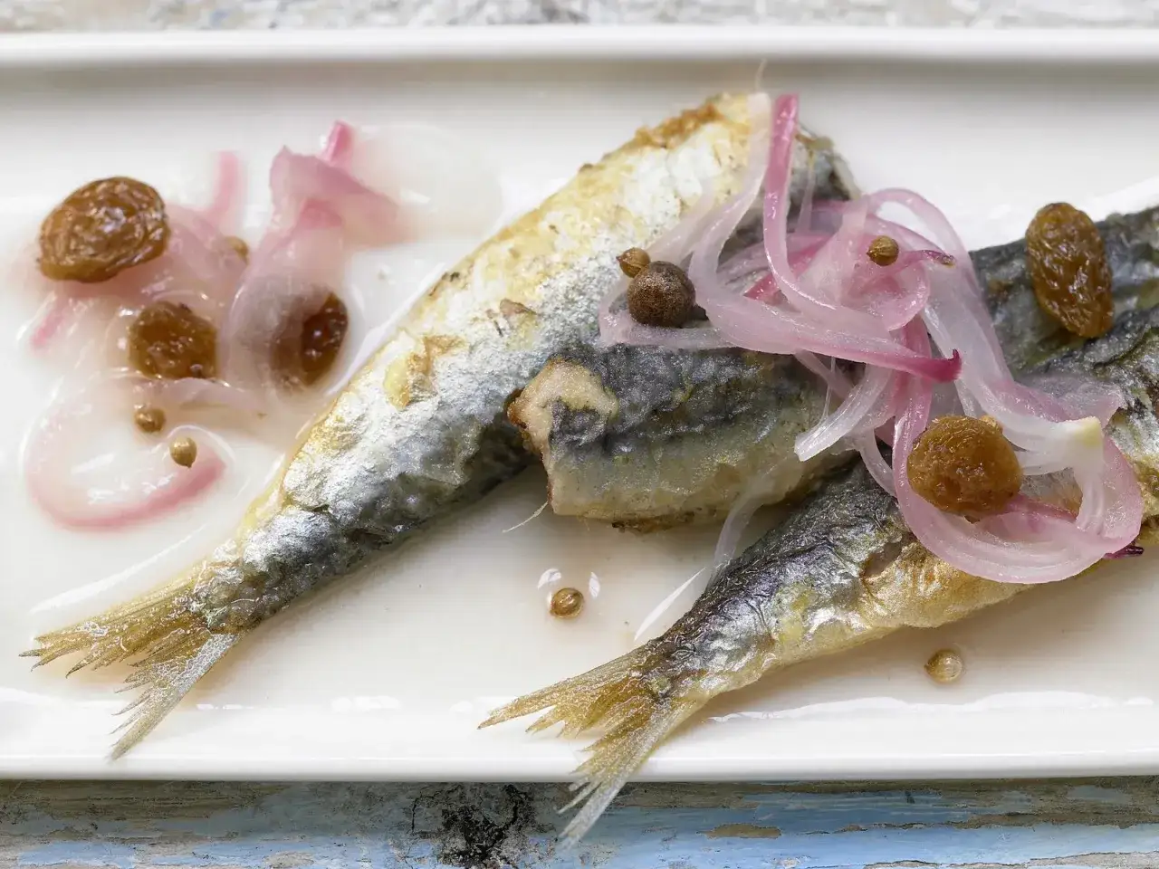 Sardinen in Saor: Gebratene Sardinen mit s&uuml;&szlig;-sauren Zwiebeln, Rosinen und Gew&uuml;rzen auf einem wei&szlig;en Teller.
