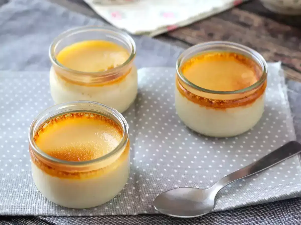 Drei Gläser mit cremigem flan caramel, die auf einem gepunkteten Tuch stehen. Eine Löffel liegt daneben.