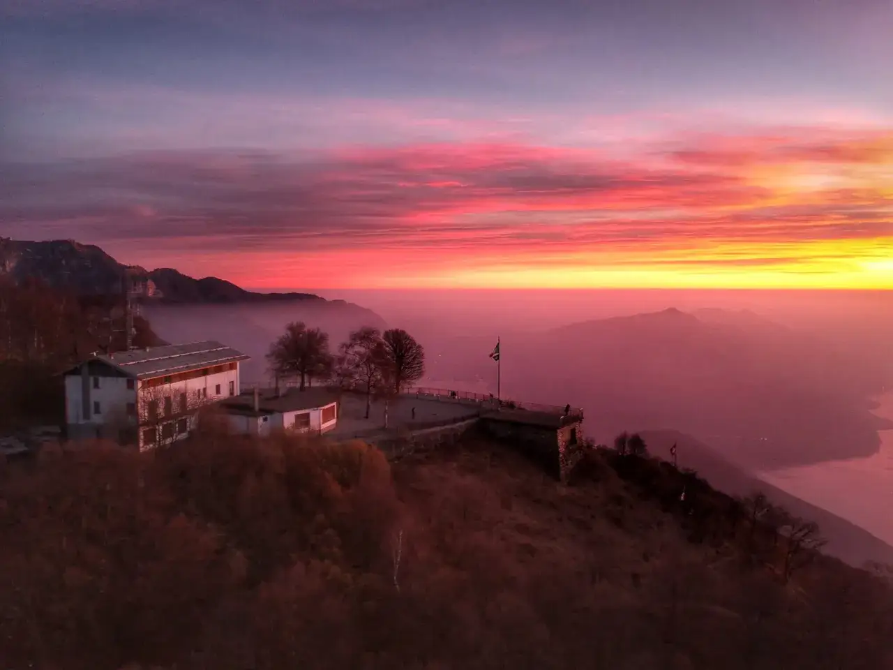 Alba infuocata sulla valle d'Intelvi, con case arroccate e un lago nebbioso all'orizzonte.