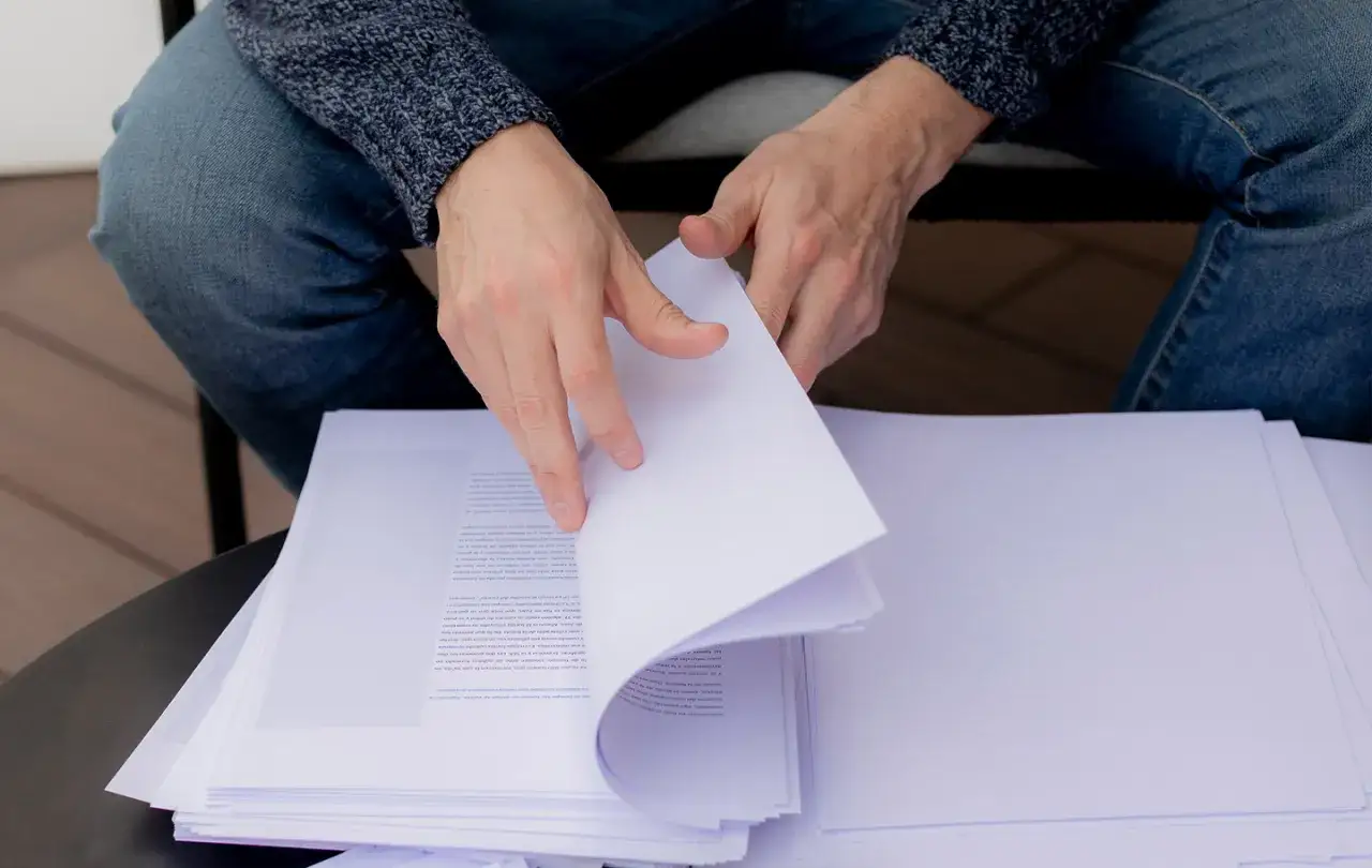 Manos revisando un mont&oacute;n de hojas impresas, prepar&aacute;ndose para el env&iacute;o de manuscritos a editoriales.