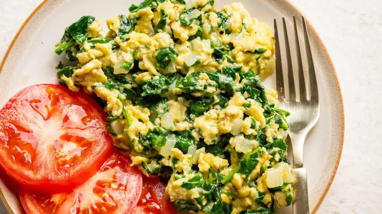 Revuelto de espinacas con trozos de cebolla, servido con rodajas de tomate fresco y un tenedor. Un desayuno saludable y delicioso.