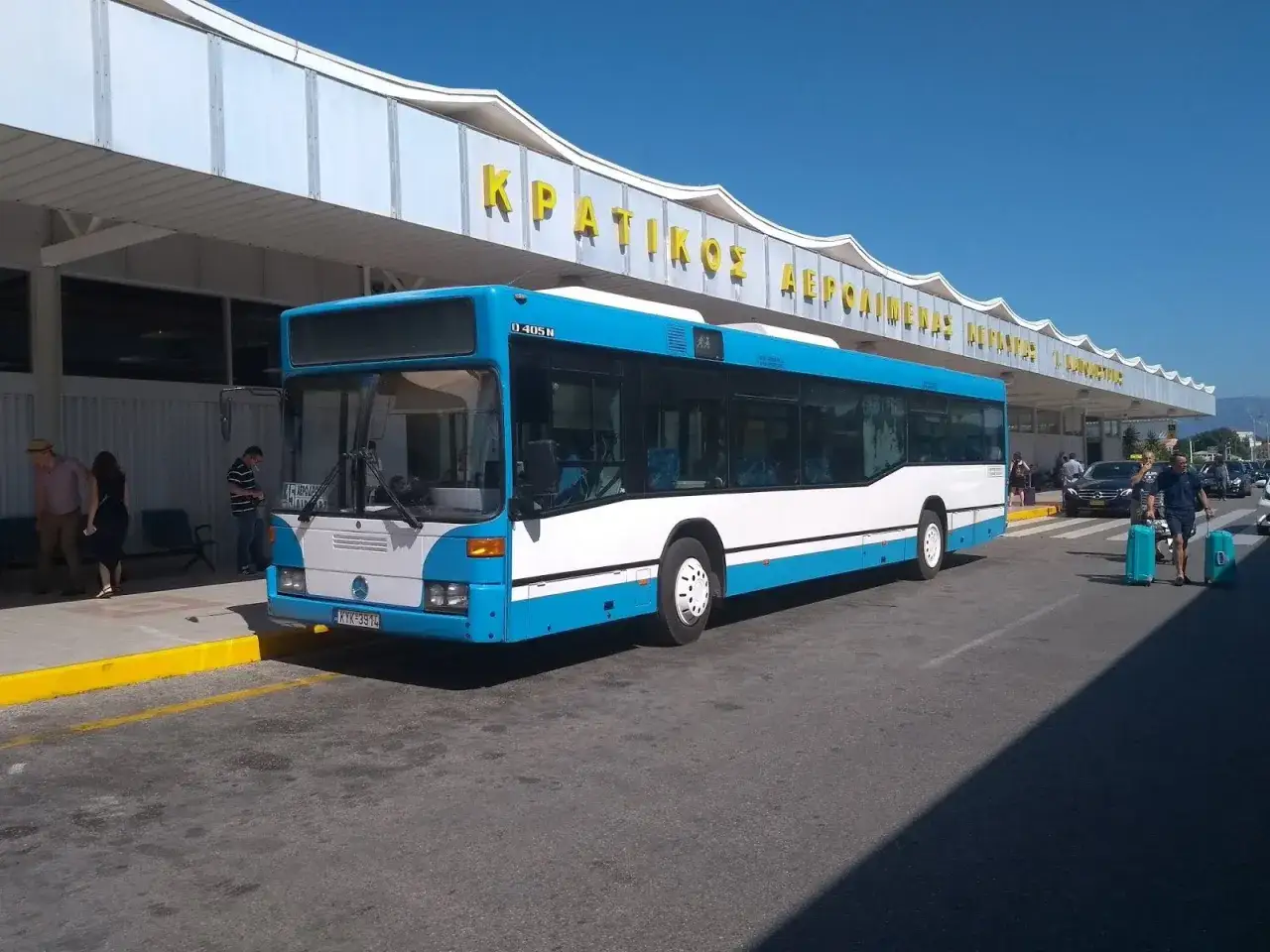 Blåvit buss vid Korfu flygplats. Soligt och folk med resväskor.