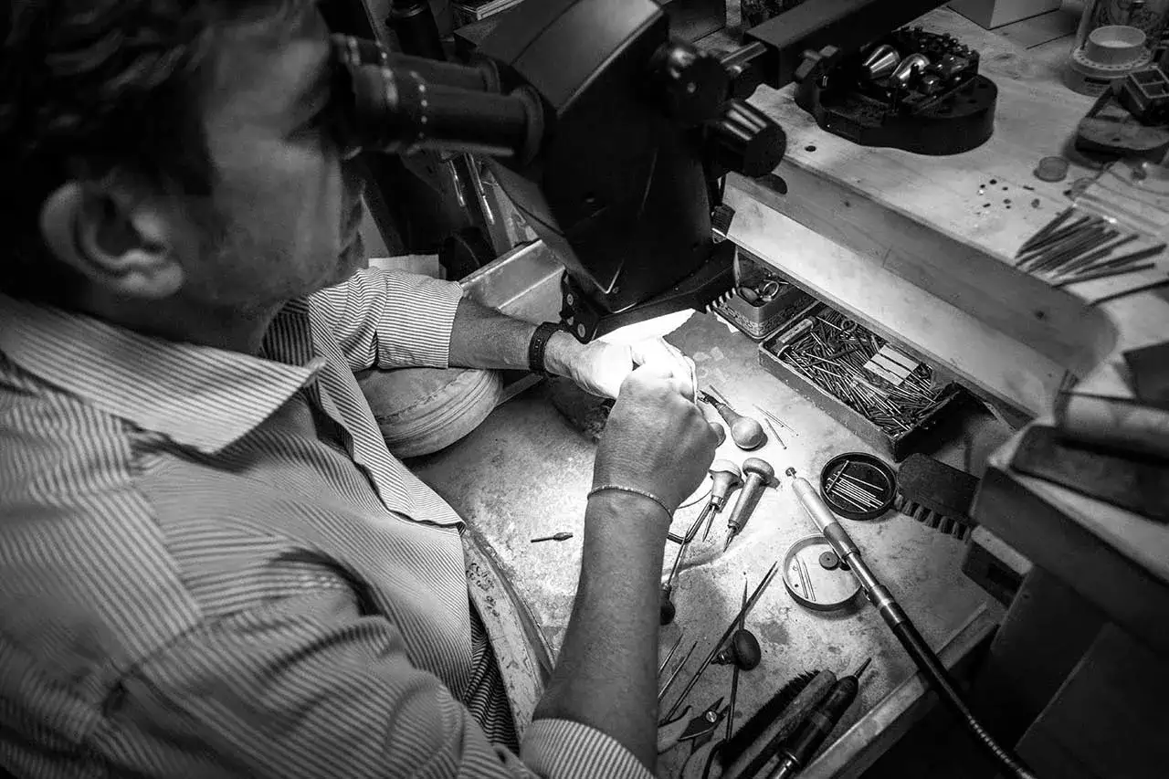 Gioielliere al microscopio, impegnato in un'incastonatura a castone ribattuto. Strumenti e gemme sul banco da lavoro.