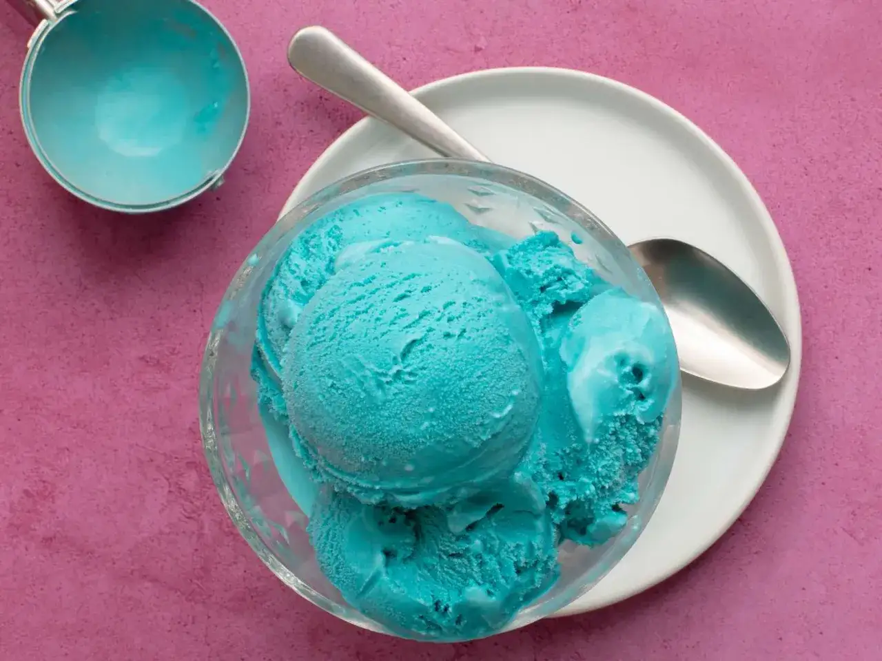 Schüssel mit leuchtend blauem Eis, daneben ein Eisportionierer und ein Löffel auf einem Teller.
