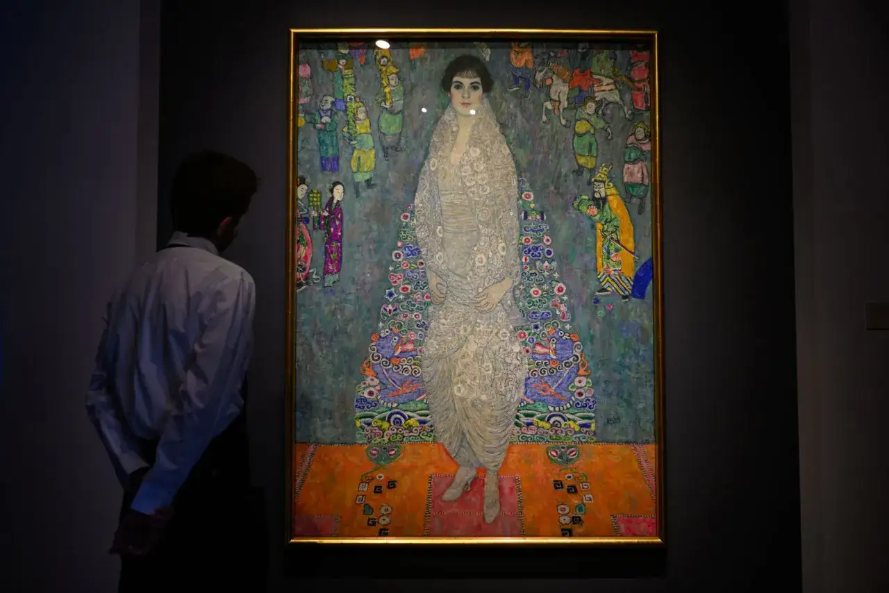 A man gazes at a Klimt painting, a masterpiece of decorative art and one of the best portraits ever created, featuring a woman in an ornate gown surrounded by vibrant figures.