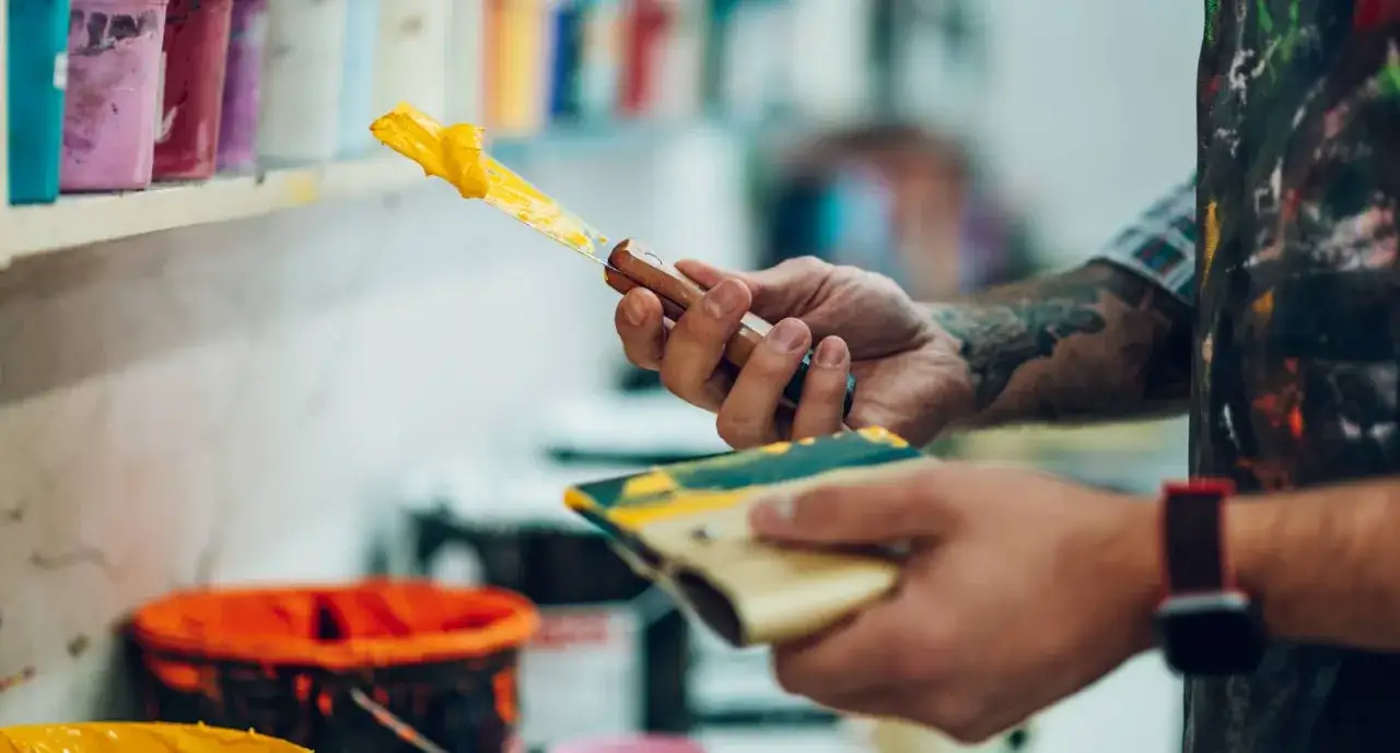 Man con tatuajes prepara pintura amarilla con espátula. Se puede pintar encima de pintura de caucho.