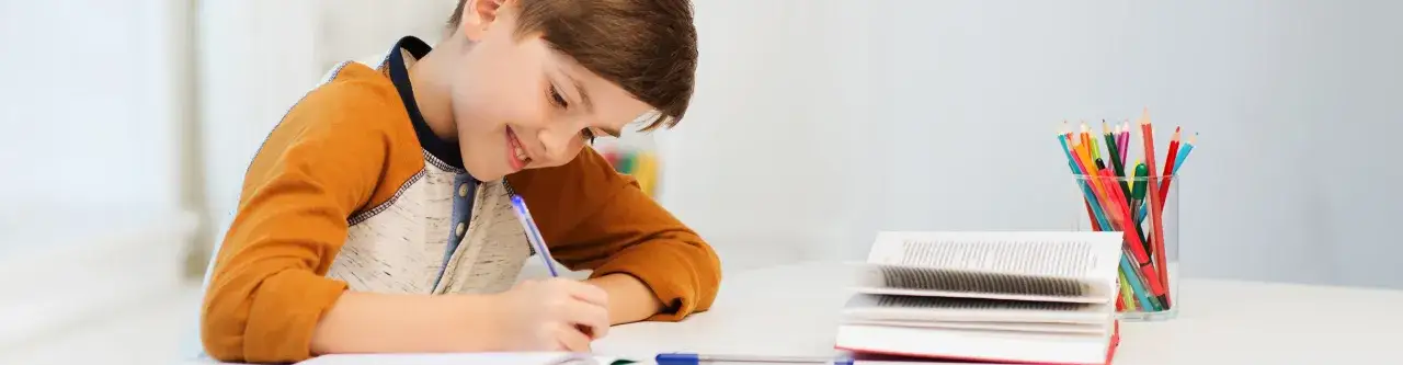 Uśmiechnięty chłopiec pisze, obok stos książek i kolorowe kredki w szklance. To jego **check lista** zadań do wykonania.