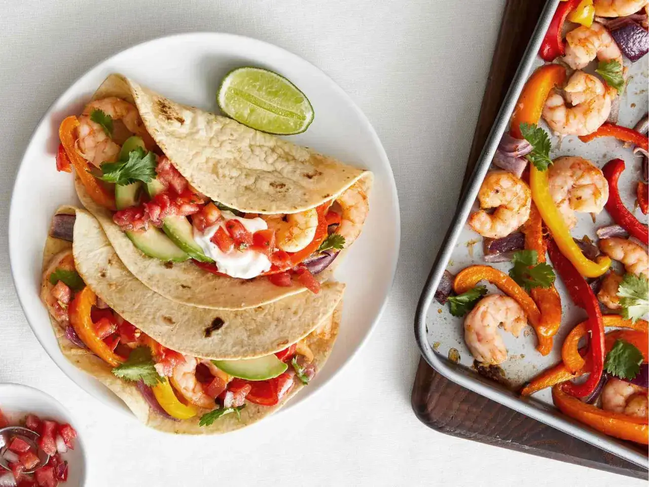 Tacos z krewetkami i warzywami to idealna lekka kolacja. Na talerzu tortille z awokado, pomidorami i krewetkami. Obok blacha z pieczonymi paprykami i krewetkami.