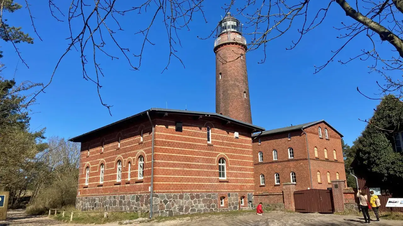Der Leuchtturm Prerow ragt imposant in den blauen Himmel. Ziegelbauten mit Rundbogenfenstern säumen seinen Fuß.