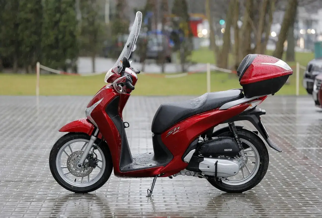 Czerwony skuter Honda SH z kuferkiem, gotowy do jazdy. Kto może jeździć skuterem? Osoby posiadające odpowiednie uprawnienia.
