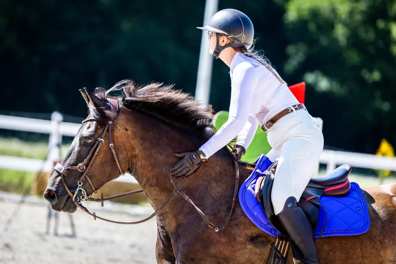 Jeździec w białym stroju i kasku siedzi na brązowym koniu. Widać siodło i niebieską podkładkę. To jakby praca saddle fittera, by zapewnić komfort.