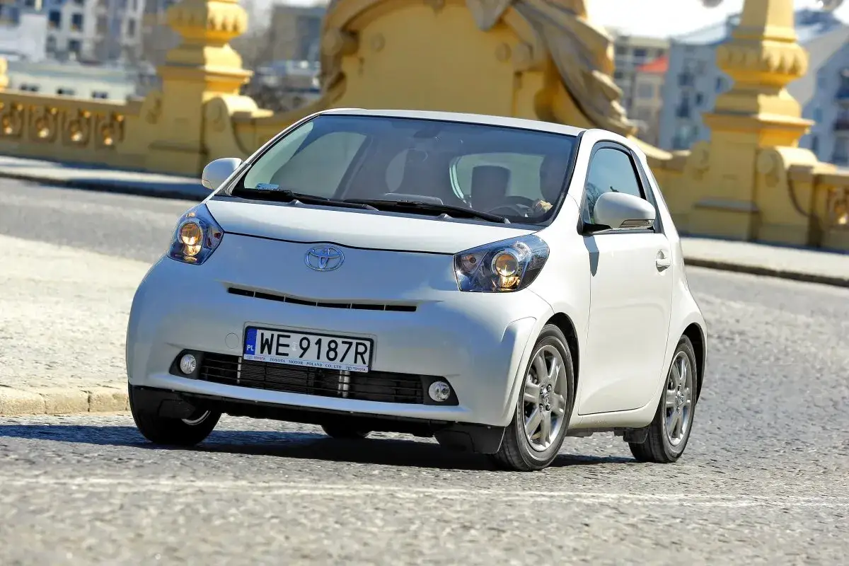 Biały, kompaktowy samochód Toyota iQ, podobny do modeli Daihatsu, jedzie po miejskiej ulicy.