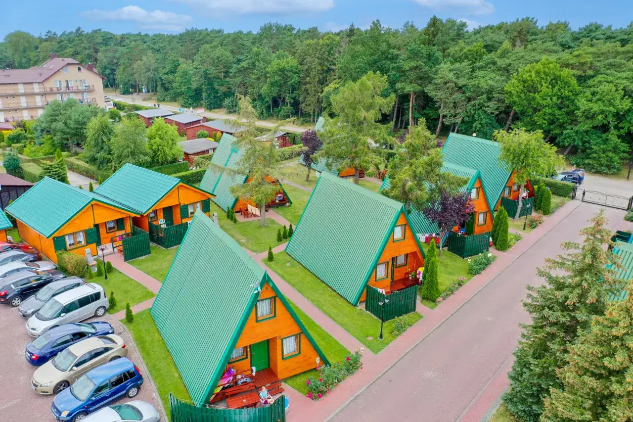 Ośrodek wczasowy Duet: drewniane domki z zielonymi dachami, otoczone drzewami, z parkingiem i budynkiem hotelowym w tle.