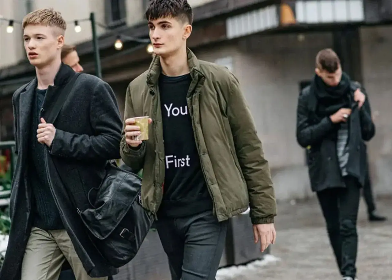Dw&oacute;ch mężczyzn w miejskim stylu, uosabiających moda skandynawska. Jeden w ciemnym płaszczu i torbie, drugi w zielonej kurtce i bluzie z napisem.