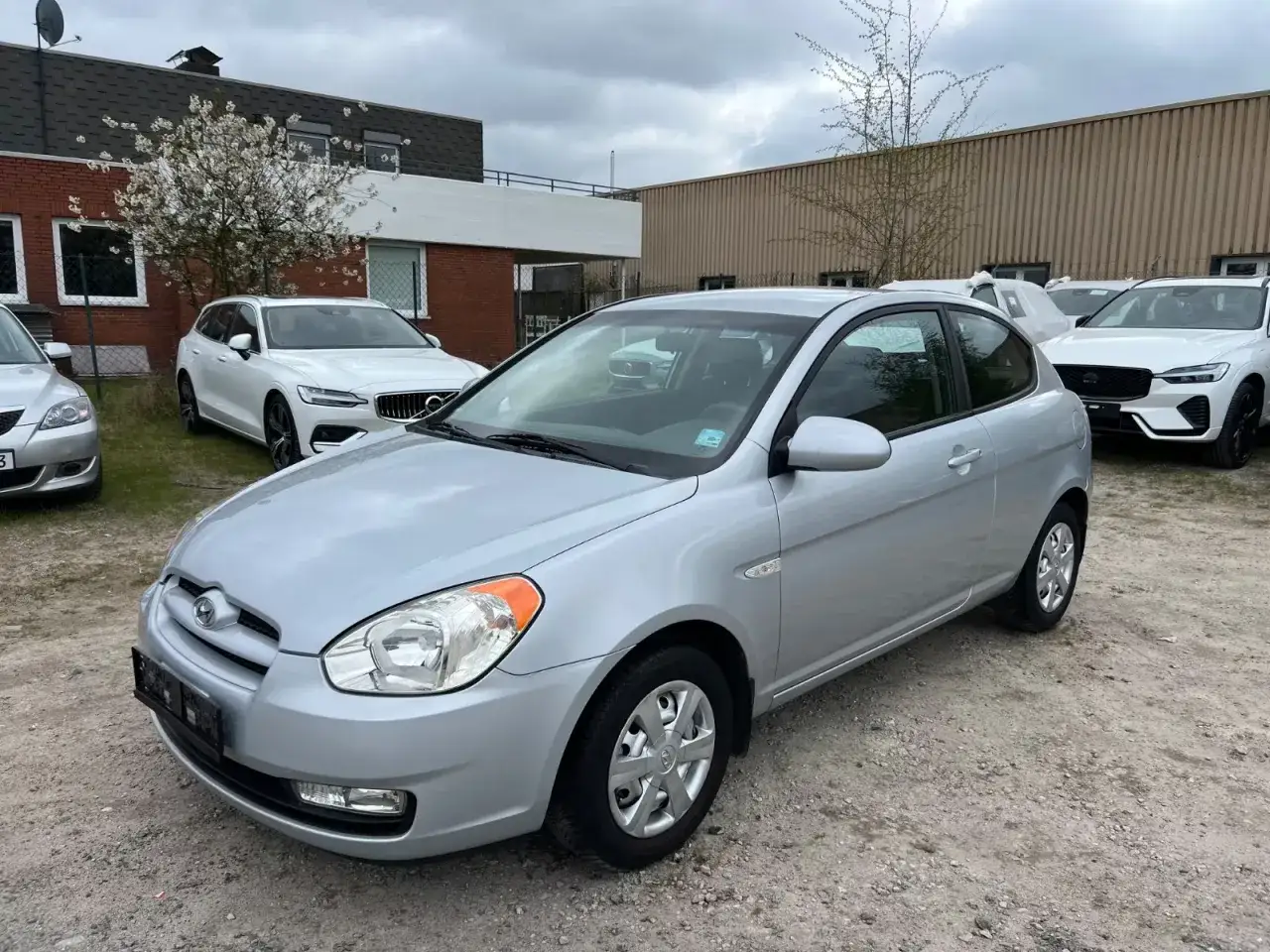 Srebrny Hyundai Accent stoi na żwirowym placu, obok białych Volvo.