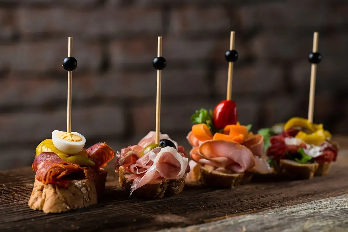 Cztery apetyczne pinchos na drewnianej desce: z jajkiem, wędlinami, warzywami i oliwkami.