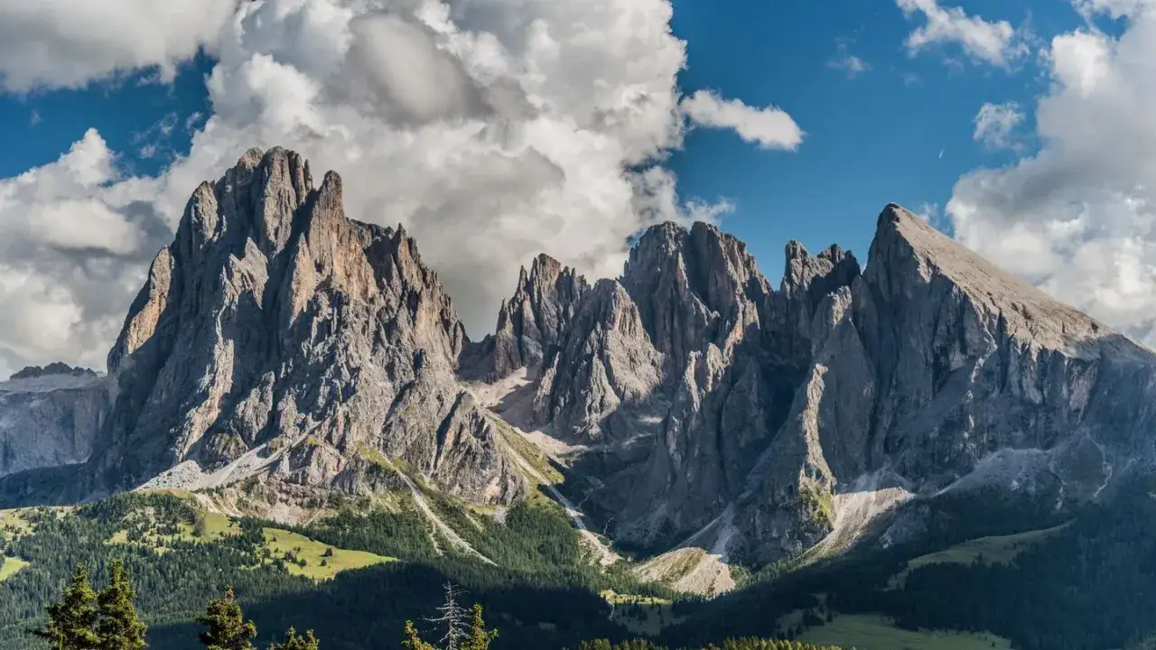Imponenti vette rocciose del Sassolungo si stagliano contro un cielo azzurro con nuvole bianche. Un sentiero serpeggia tra i verdi prati e i boschi sottostanti.