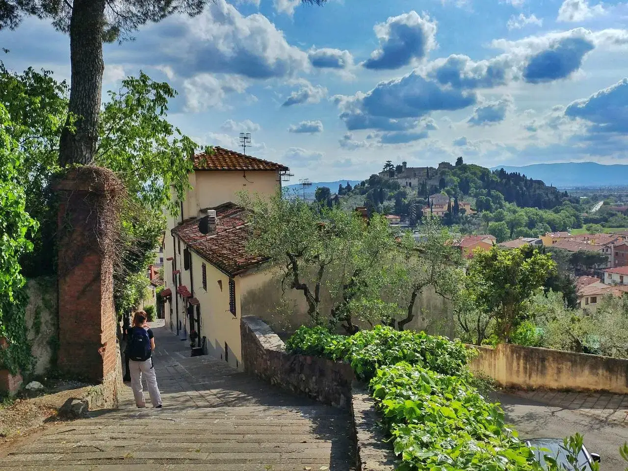 Una persona scende una scalinata in pietra verso un borgo con vista su **Monte Morello** e il suo castello, sotto un cielo azzurro con nuvole bianche.
