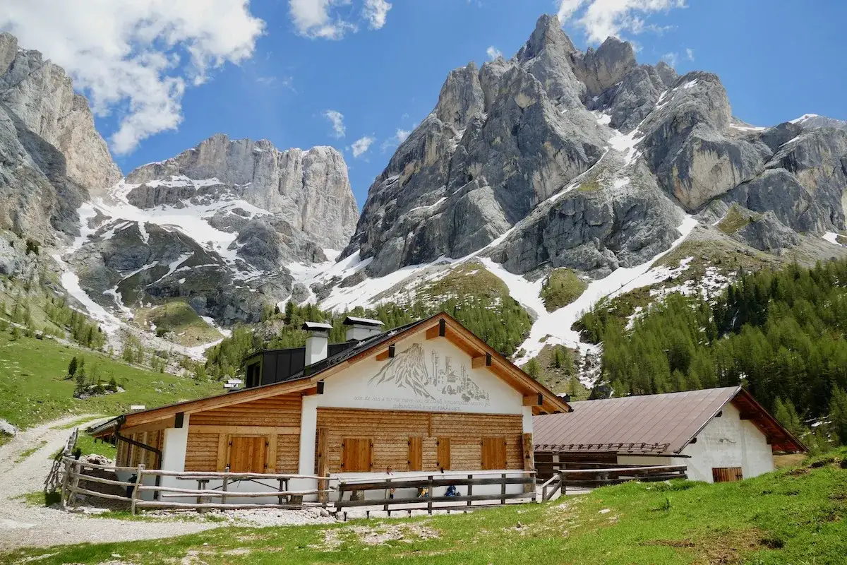 Malga Contrin, un rifugio alpino in legno con tetto scuro, si staglia contro imponenti montagne rocciose innevate e un cielo azzurro con nuvole bianche.