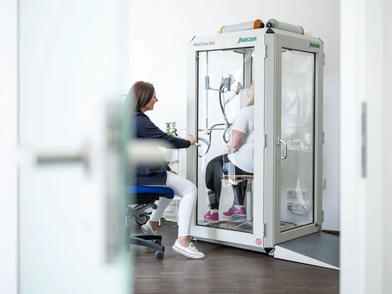 Ärztin überwacht Lungenfunktionstest in einer Ganzkörperkammer. Nach Lungenlappenentfernung kann dies zur Beurteilung einer Schwerbehinderung dienen.