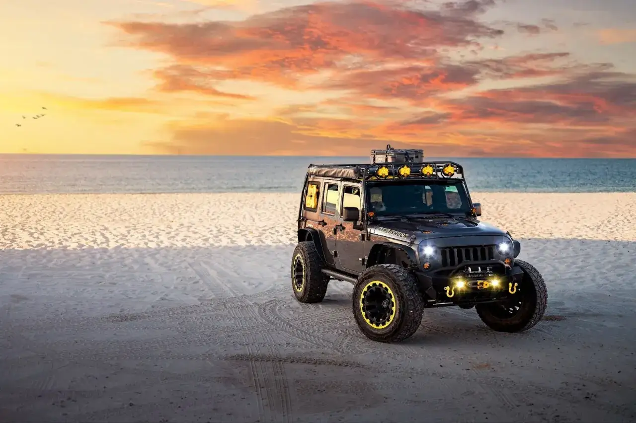 Czarny Jeep na plaży o zachodzie słońca, gotowy na przygodę. To jeden z najlepszych samochodów terenowych, który poradzi sobie w każdym terenie.