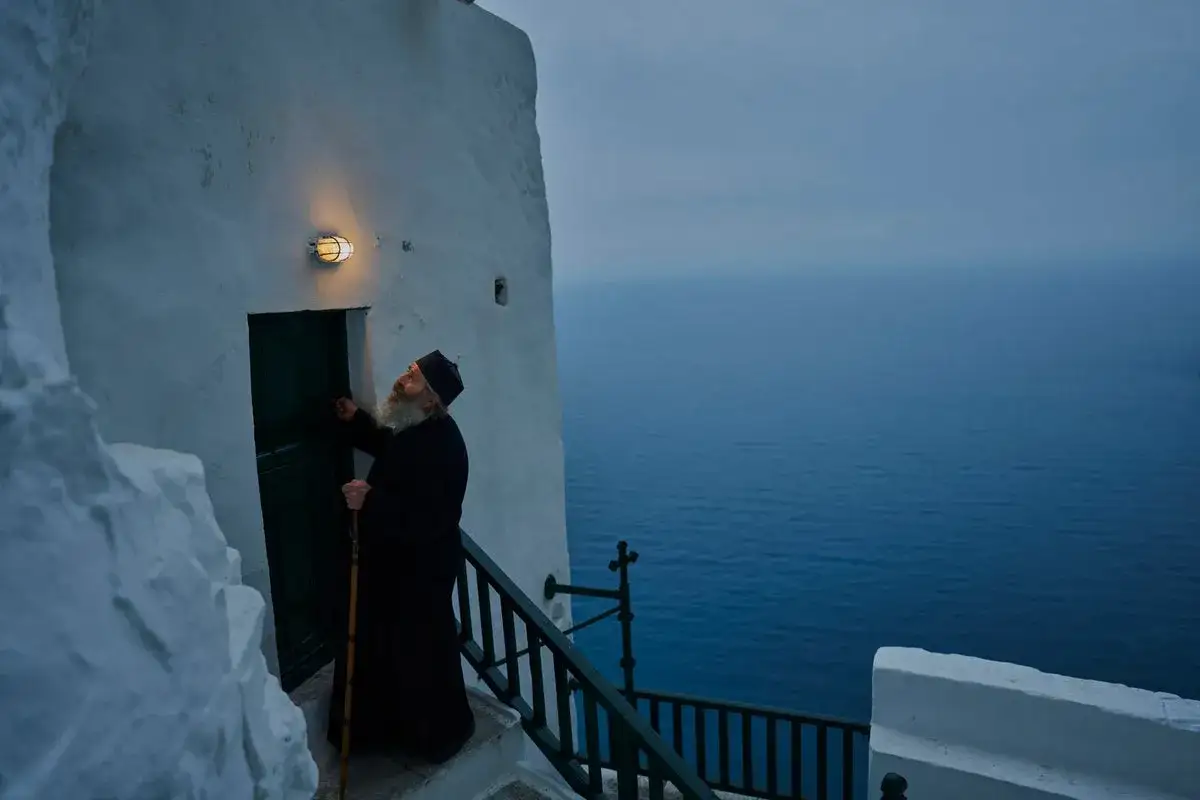 A bearded monk in black robes stands on a staircase, knocking on a door. The vast blue sea stretches out behind him, a serene backdrop for this moment of contemplation.