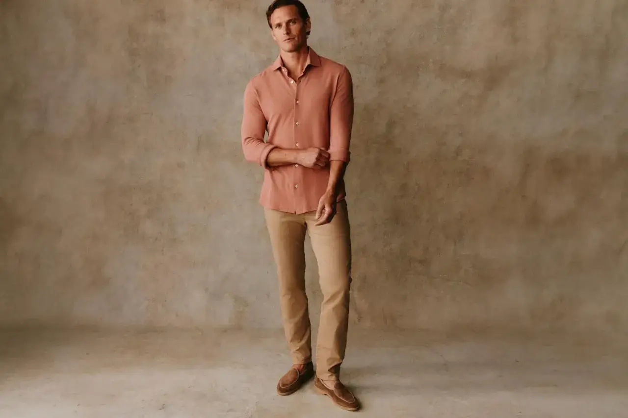 A man models a salmon-colored shirt and tan chino pants, showcasing a casual yet refined look.