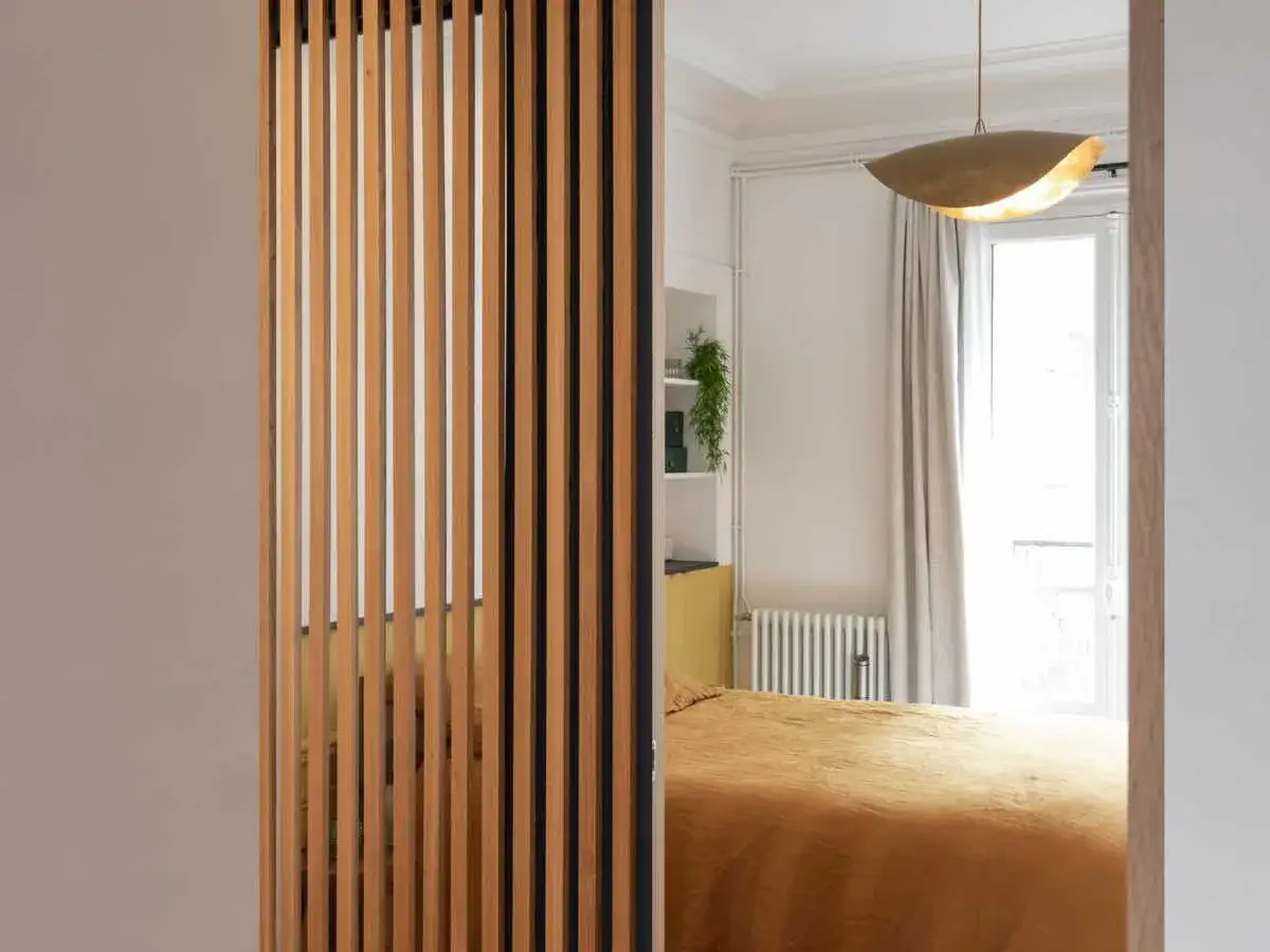Un séparateur de chambre en bois crée une division élégante, révélant un lit douillet et une fenêtre lumineuse.