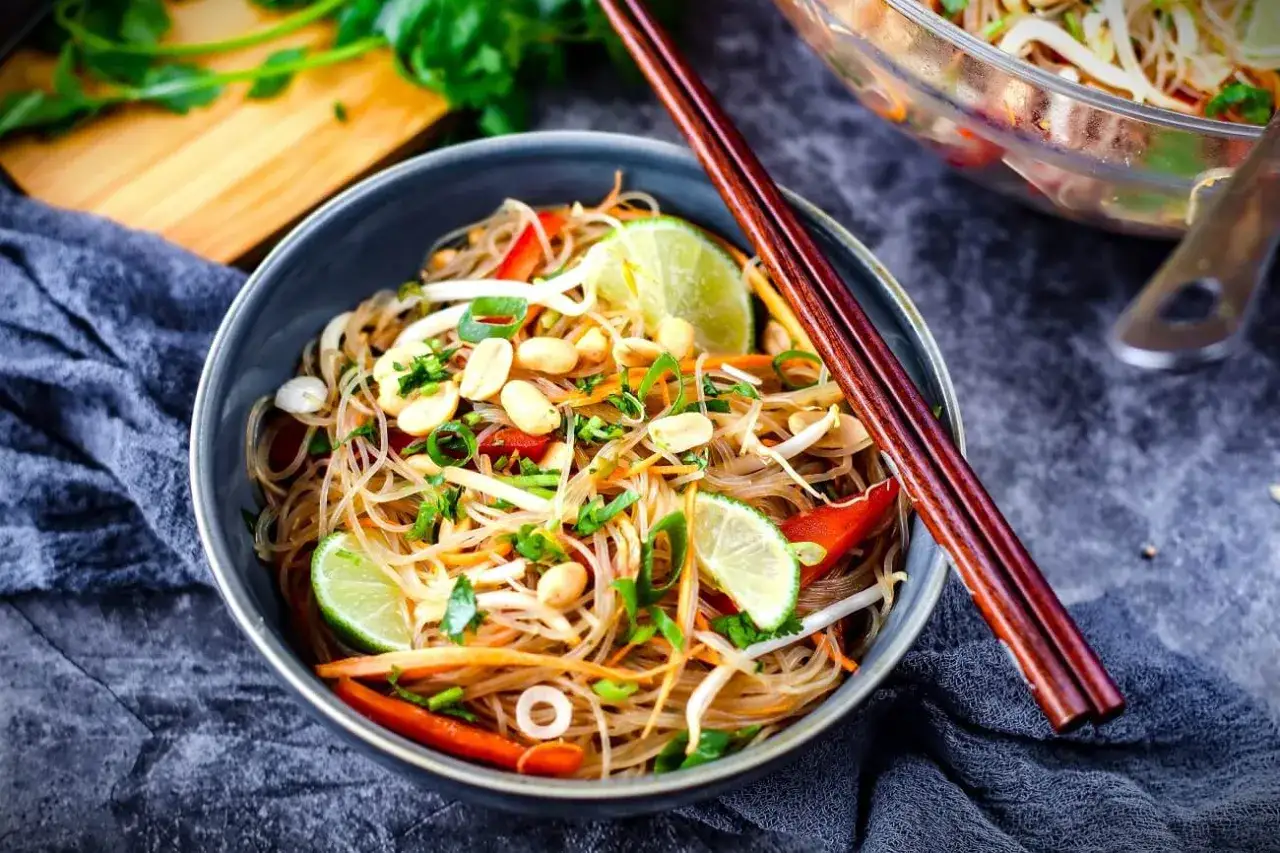 Sch&uuml;ssel mit Glasnudelsalat, garniert mit Erdn&uuml;ssen, Limettenspalten und Koriander. Chopsticks liegen auf der Sch&uuml;ssel.