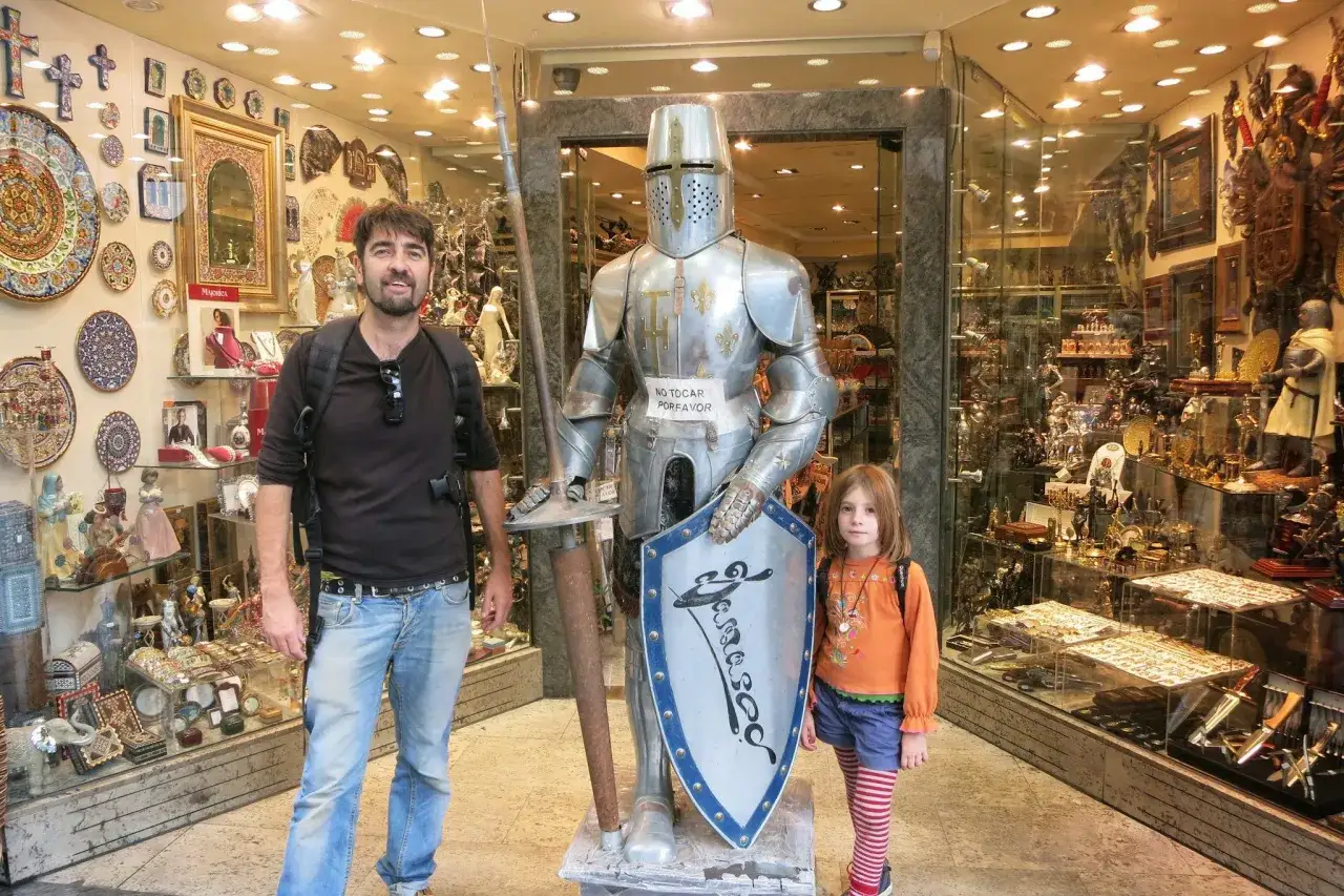 Un padre y su hija posan junto a una armadura de caballero en una tienda de recuerdos en Toledo con niños.