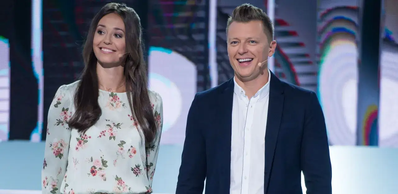 Uśmiechnięta para prowadzi Koło Fortuny. Ona w kwiecistej sukience, on w granatowej marynarce.
