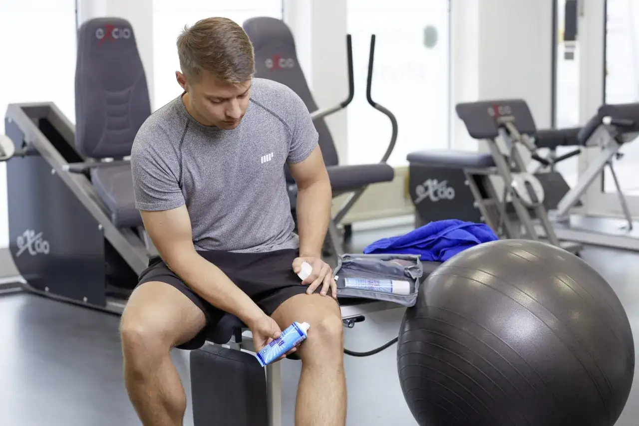 Mann im Fitnessstudio tr&auml;gt Salbe auf sein Knie auf. Was hilft bei Muskelschmerzen? Ein Sportler mit Schmerzsalbe und Gymnastikball.