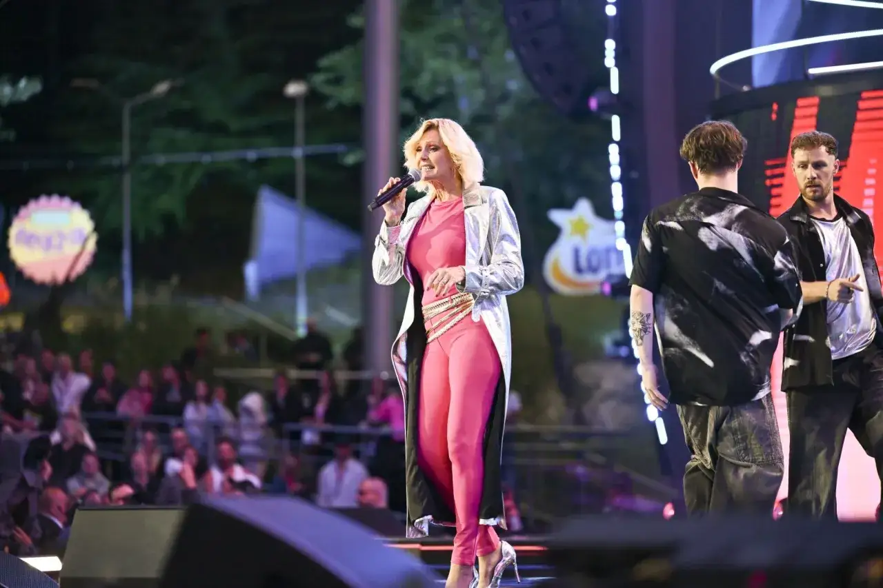 Helena Vondráčková w różowym stroju i srebrnym płaszczu śpiewa na scenie, podczas gdy dwaj mężczyźni stoją obok niej.