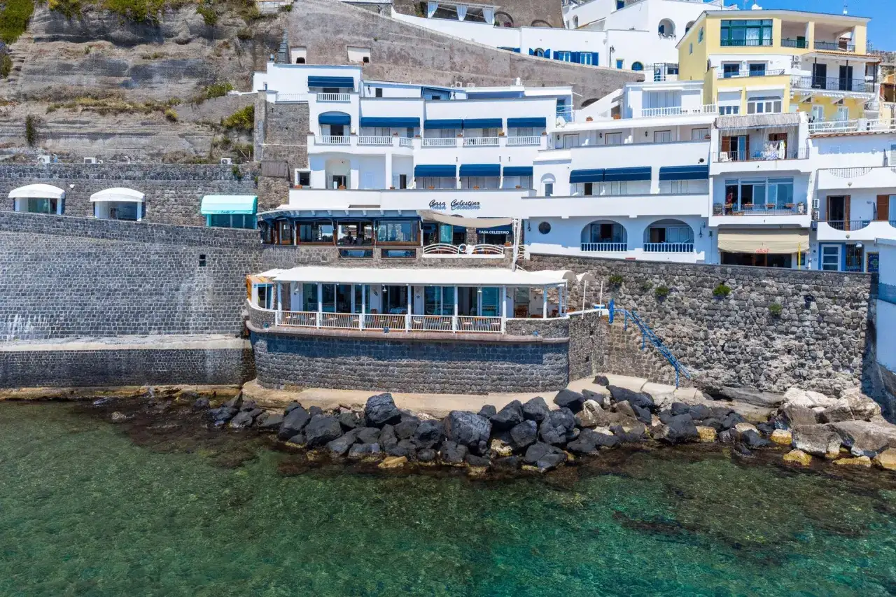 Hotel Celestino Ischia, con la sua architettura bianca e blu, si affaccia sul mare cristallino, incastonato tra rocce e vegetazione.