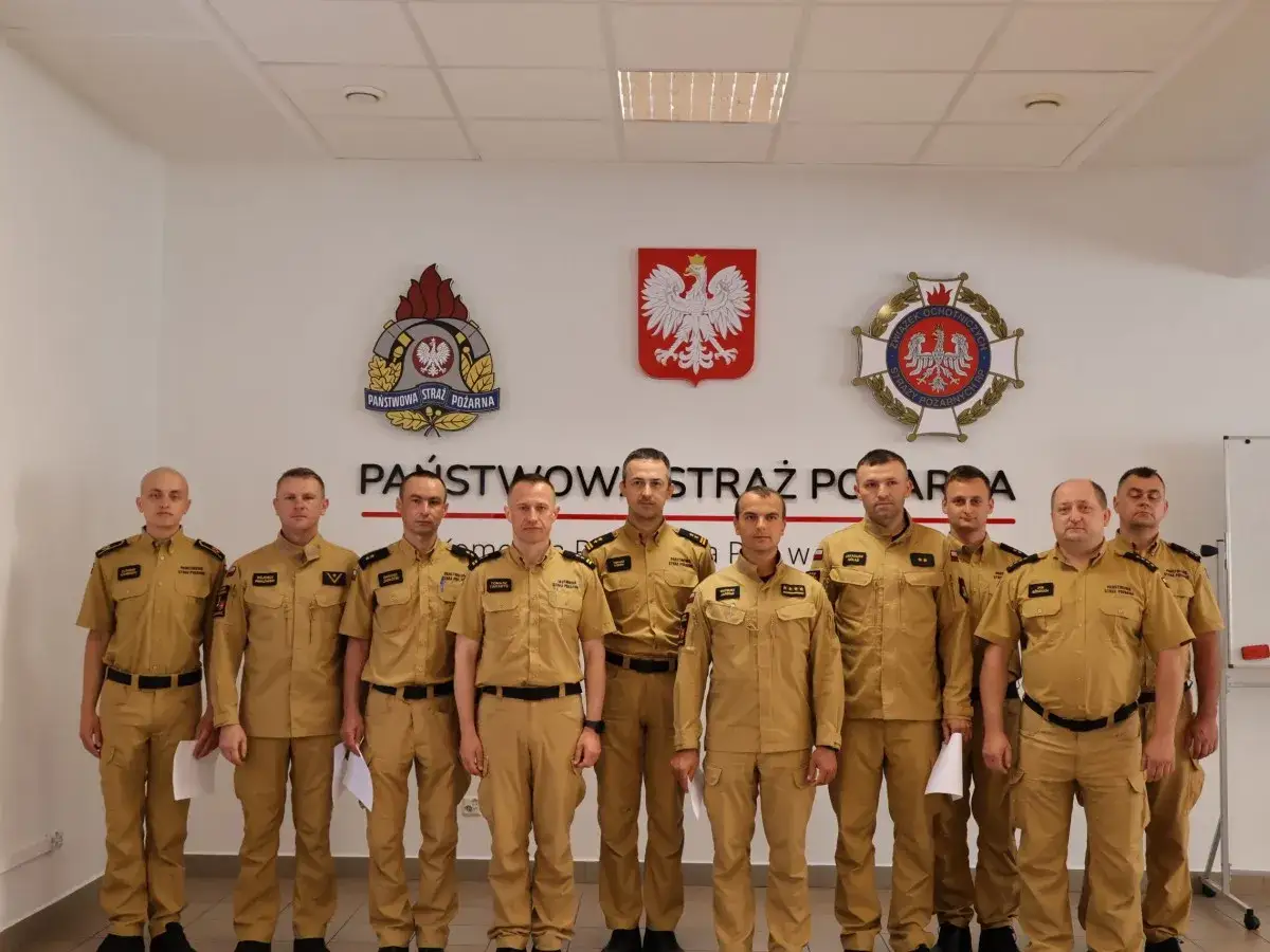 Grupa strażaków w beżowych mundurach, wśród nich brygadier, pozuje do zdjęcia na tle godła Państwowej Straży Pożarnej.