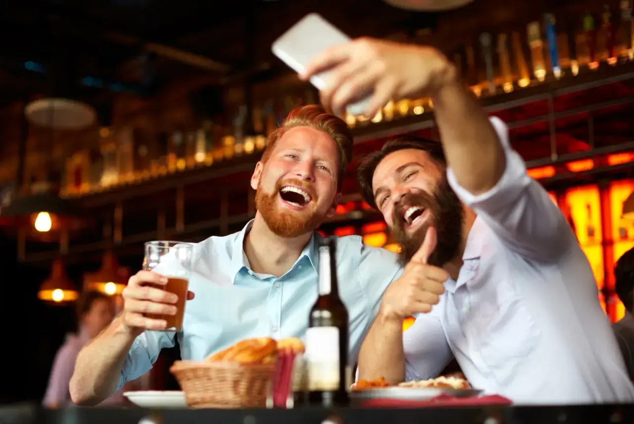 Dos clientes de restaurante riendo y tom&aacute;ndose un selfie con cerveza y comida.