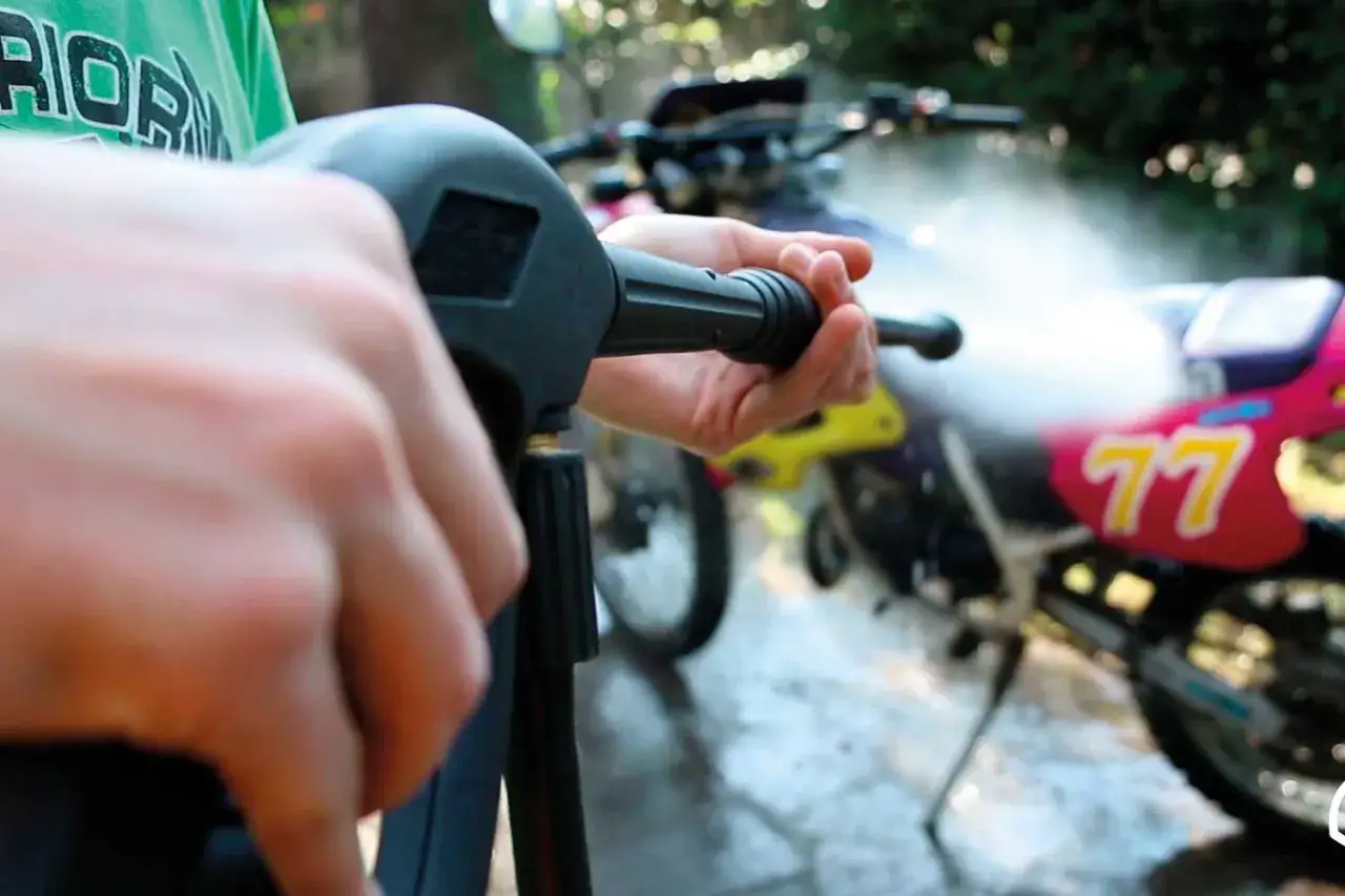 Mani che impugnano un'idropulitrice per mostrare come lavare la moto a casa. L'acqua schizza sulla moto rosa e gialla.