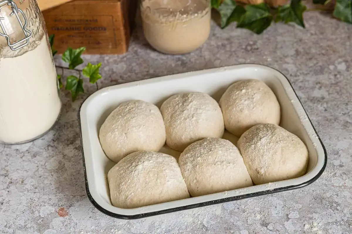 Six boules de p&acirc;te &agrave; pain, saupoudr&eacute;es de farine, reposent dans un plat blanc. &Agrave; c&ocirc;t&eacute;, un bocal contient mon levain, pr&ecirc;t &agrave; faire lever le pain.