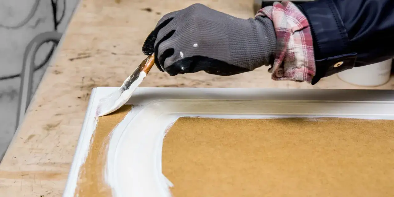 Mano enguantada pintando un marco de mdf con brocha blanca.