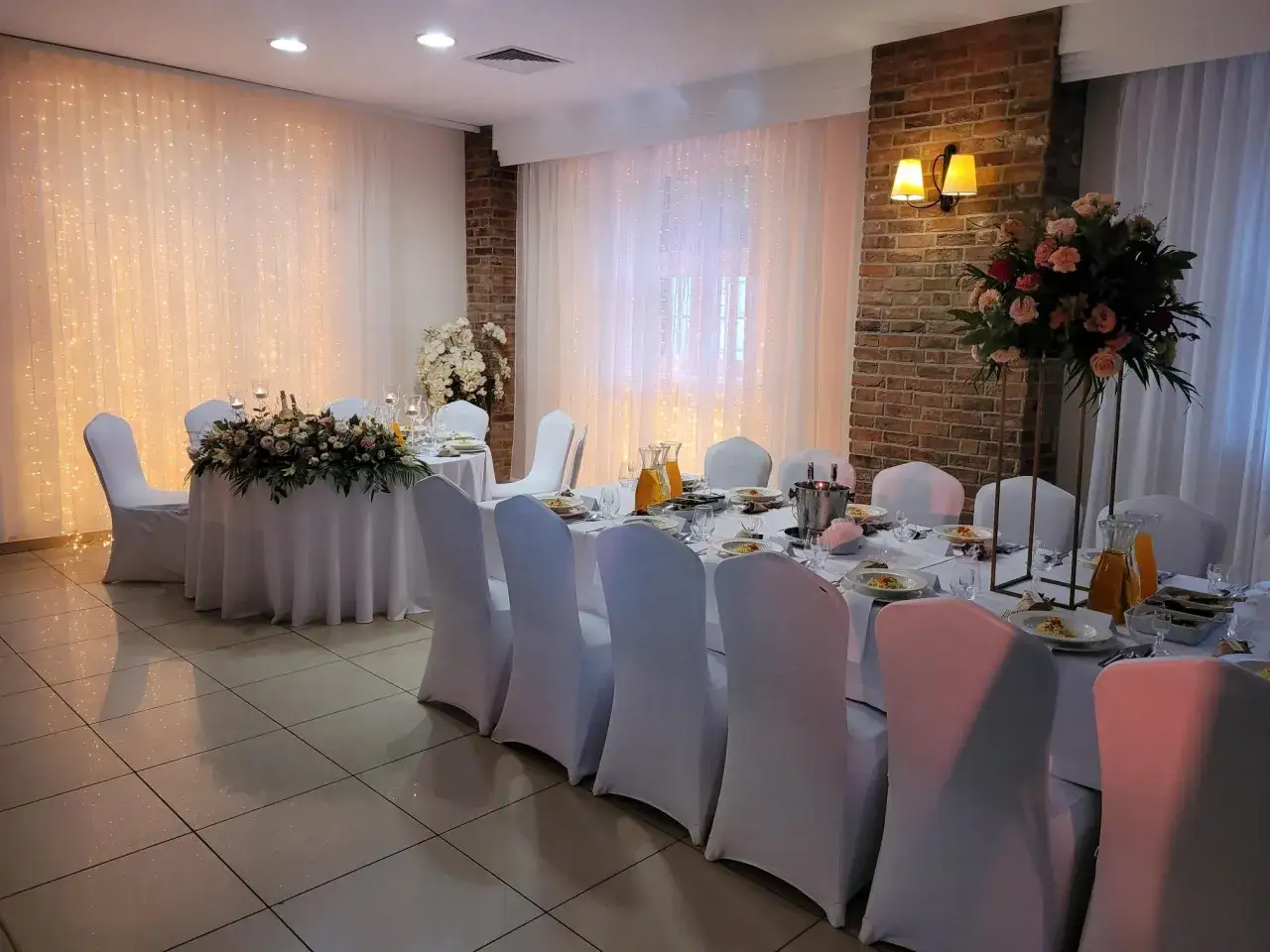 Elegancka restauracja przygotowana na wesele. Stoły nakryte białymi obrusami, ozdobione kwiatami i świecami. W tle migoczące światełka.